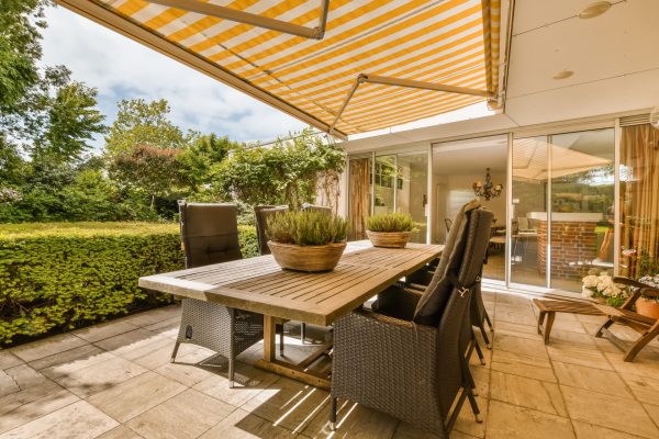 an outdoor dining area with table and chairs under the awning on a sunny day in the patio is surrounded by lush green trees